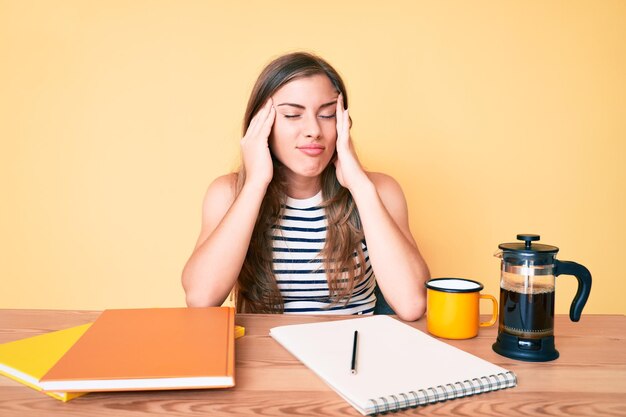 Mooie jonge blanke vrouw zit op de tafel te studeren voor de universiteit met hand op hoofdpijn omdat stress lijdt aan migraine