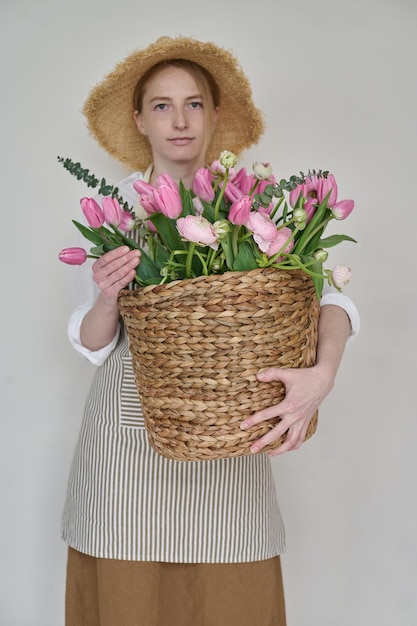 Mooie jonge bloemist met een rieten mand met bloemen in haar handen en een strohoed op een witte achtergrond