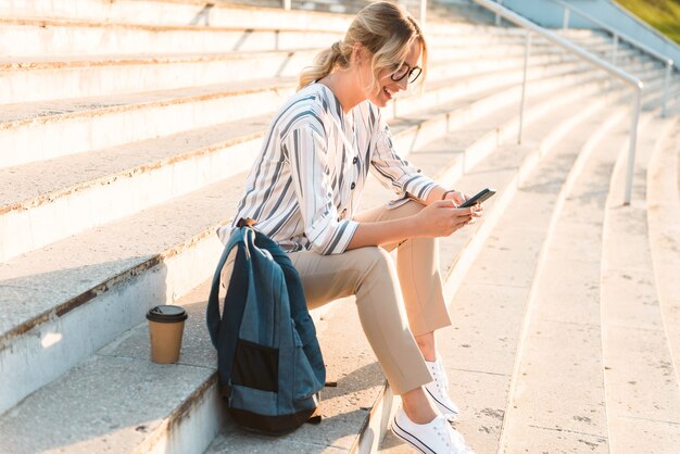 Mooie jonge blonde meid die mobiele telefoon gebruikt terwijl ze buiten op trappen zit en koffie drinkt