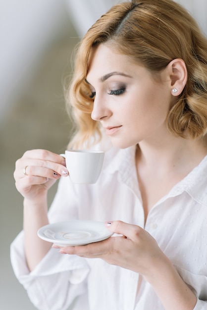 Mooie jonge blonde meisje in een licht shirt in een lichte kamer geniet van een kopje koffie in de ochtend voordat ze naar het werk gaat. Meisje in de badkamer in openhartige kleding.