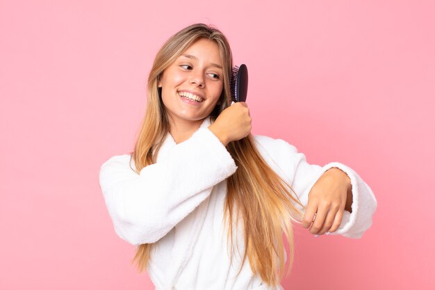 Mooie jonge blonde vrouw die een badjas draagt en een haarborstel vasthoudt