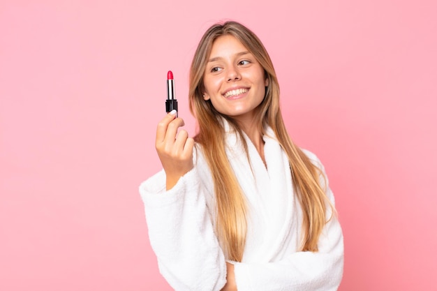 Mooie jonge blonde vrouw die een badjas draagt en een lippenstift vasthoudt