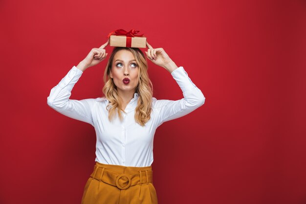 Mooie jonge blonde vrouw geïsoleerd op rode achtergrond, met geschenkdoos op haar hoofd