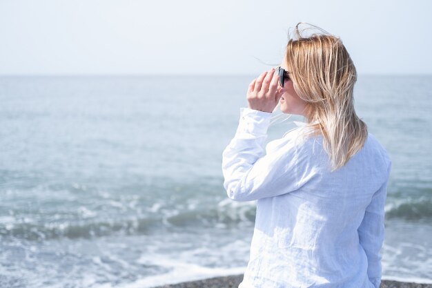 Mooie jonge blonde vrouw in wit overhemd en zonnebril staande aan de kust van de zee