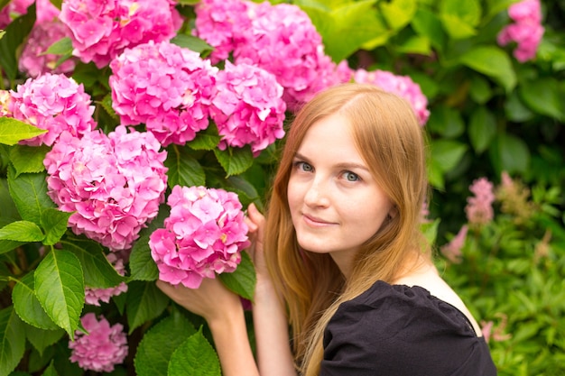 Mooie jonge blonde vrouw met bloemen in de buurt van gezicht. Roze Hydrangea-macrophylla die in de zomer in botanische tuin bloeien