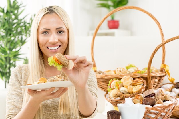 Mooie jonge blonde vrouw met een glimlach op haar gezicht die naar de camera kijkt, een mini-sandwich vasthoudt en de kijker een beetje aanbiedt. Op de achtergrond zie je een geweven mand met bakkerijproducten.