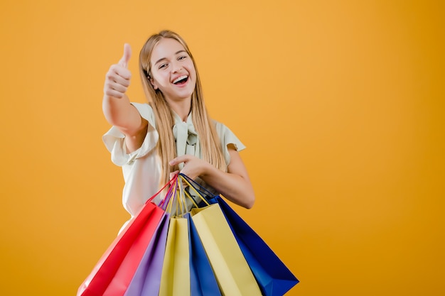 Mooie jonge blondevrouw die met kleurrijke die het winkelen zakken glimlachen over geel worden geïsoleerd