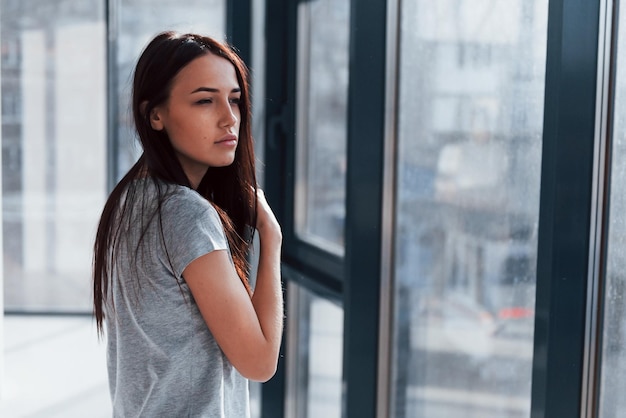 Mooie jonge brunette in grijs shirt staande in de buurt van raam en poseren voor een camera.