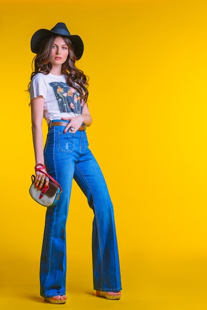 Mooie jonge brunette meisje in witte t-shirt spijkerbroek uitlopende met een kleine tas en een hoed staande op een gele achtergrond