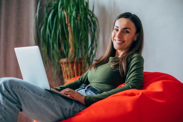 Mooie jonge brunette vrouw die thuis laptop gebruikt terwijl ze op een stoel zit glimlachend Freelance levensstijl concept rust ontspannen