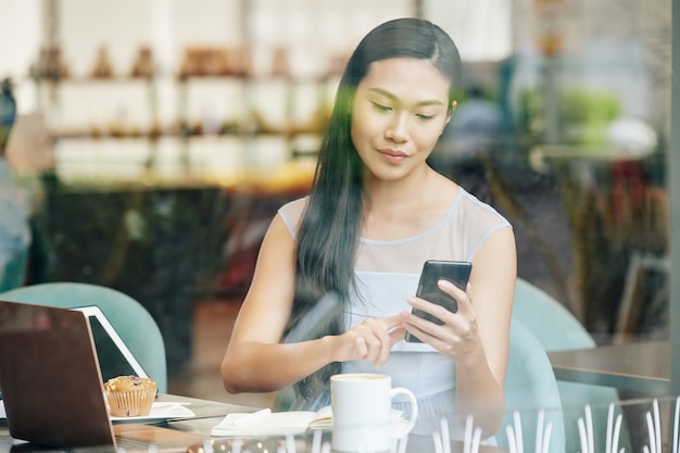 Mooie jonge Chinese vrouw die aan tafel in het café werkt en tekstberichten in smartphone controleert