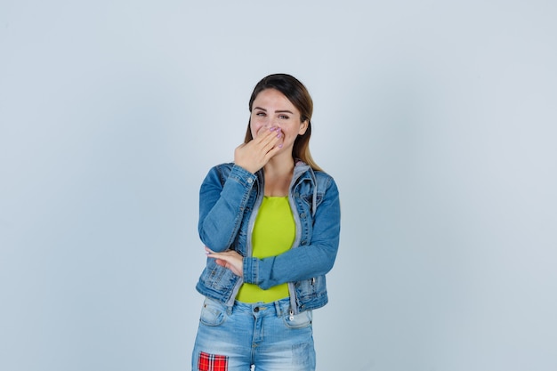 Mooie jonge dame die de hand op de mond houdt in denim outfit en er vrolijk uitziet, vooraanzicht.