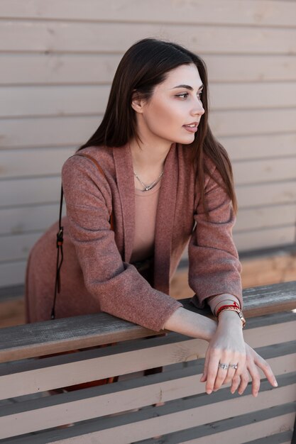 Mooie jonge elegante vrouw mannequin in modieuze elegante jas rust in de buurt van houten gebouw in de stad. Mooi stijlvol aantrekkelijk meisje in jeugdkleren staat in de buurt van de muur van planken in de straat.