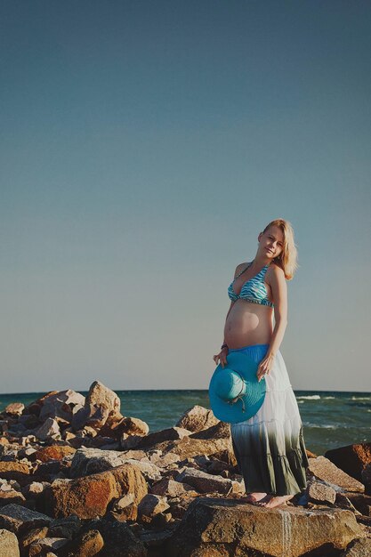 Mooie jonge elegante zwangere blonde vrouw in een blauwe hoed loopt op het strand, kust bij zonsondergang.