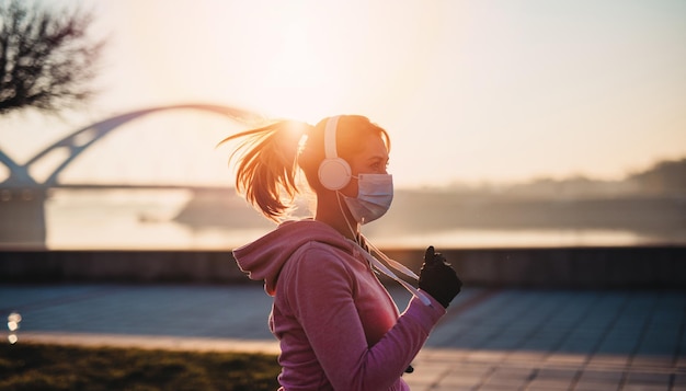 Mooie jonge en fitte vrouw in goede vorm rennen en joggen alleen op City Bridge Street. Ze draagt een beschermend gezichtsmasker om zichzelf te beschermen tegen een virus- of allergie-infectie. Zonsondergang op de achtergrond.