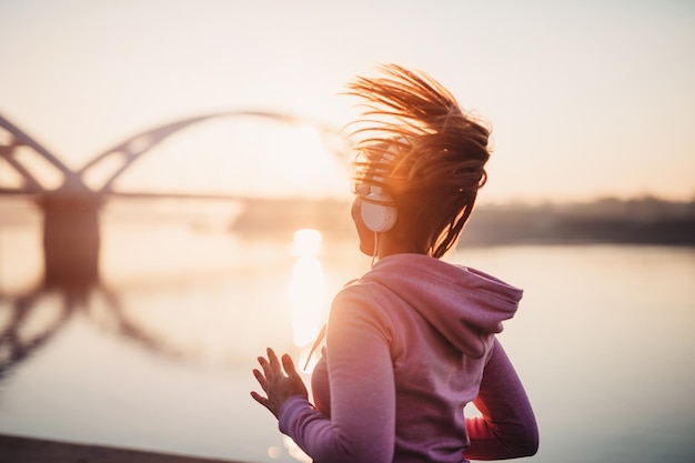 Mooie jonge en fitte vrouw in goede vorm rennen en joggen alleen op City Bridge Street. Ze luistert naar muziek met een koptelefoon. Prachtige zonsondergang op de achtergrond.