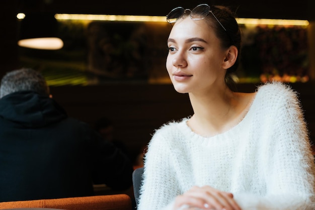 Mooie jonge freelancer meisje in witte trui zit in café glimlachend en denkend aan werk