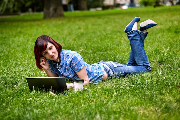 Mooie jonge freelancervrouw die laptop met behulp van die op het gras in het park liggen.