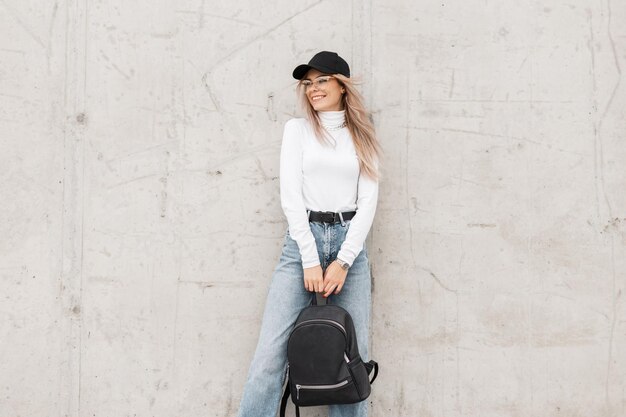 Mooie jonge gelukkige meid met een glimlach die modieuze kleding draagt met een pet, witte trui, jeans en een rugzak staat in de buurt van een betonnen grijze muur