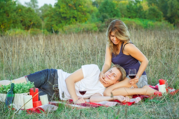 Mooie jonge gelukkige verliefde paar op picknick liggen op plaid in veld op zonnige zomerdag genieten, vasthouden en drinken van wijn en rusten. camera kijken en glimlachen.