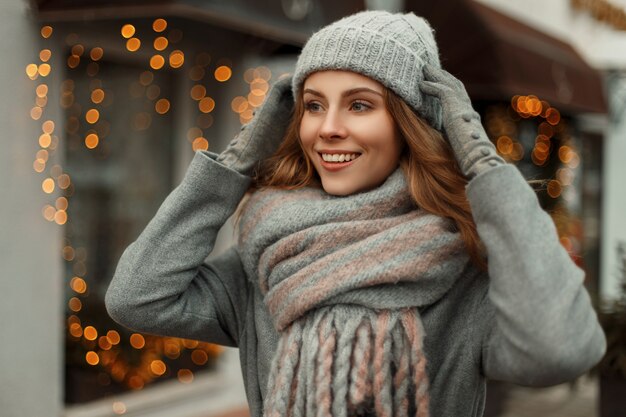 Mooie jonge gelukkige vrouw die lacht in een grijze jas met een trendy gebreide muts en stijlvolle mode sjaal wandelen in de stad op vakantie