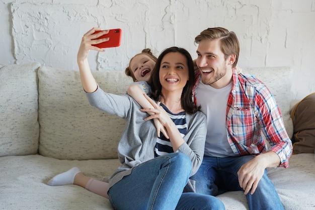 Mooie jonge gezin met klein kind dat een selfie maakt met een smartphone thuis op de bank.