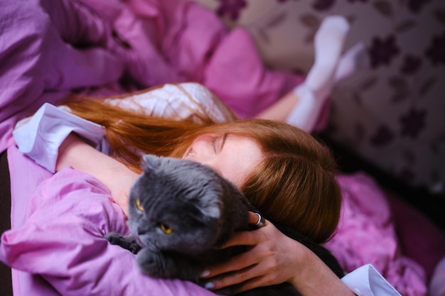 mooie jonge ginger vrouw met haar grijze Britse kat in roze bed thuis meisje in wit shirt knuffelen de kat