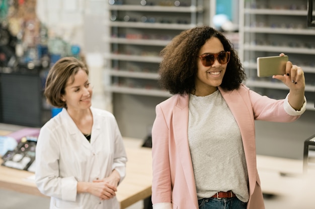 Mooie jonge glimlachende cliënt van optica winkel nieuwe zonnebril proberen en selfie maken met adviseur