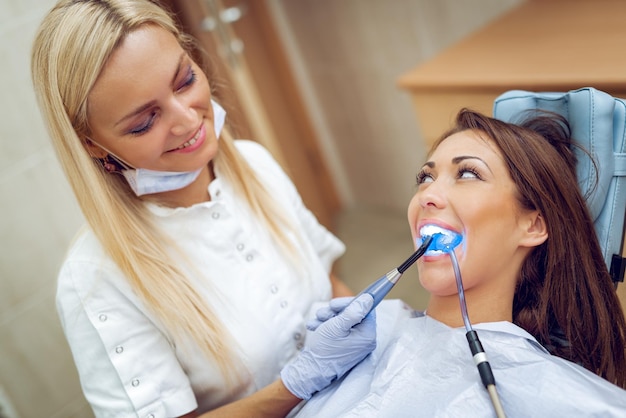 Foto mooie jonge glimlachende vrouw bij bezoek in het tandartskantoor en tandarts die de reparatietand afwerkt met tandheelkundige uithardende uv-lamp.