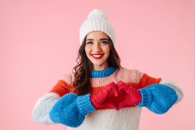 Mooie jonge glimlachende vrouw die sweater en een hoed draagt die zich geïsoleerd over roze bevindt