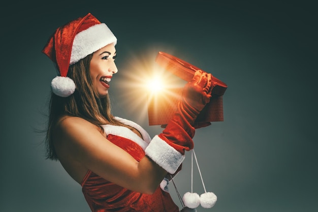 Mooie jonge glimlachende vrouw in het kostuum van de Kerstman die een rood cadeau vasthoudt en plezier heeft met kijken naar wat erin zit.