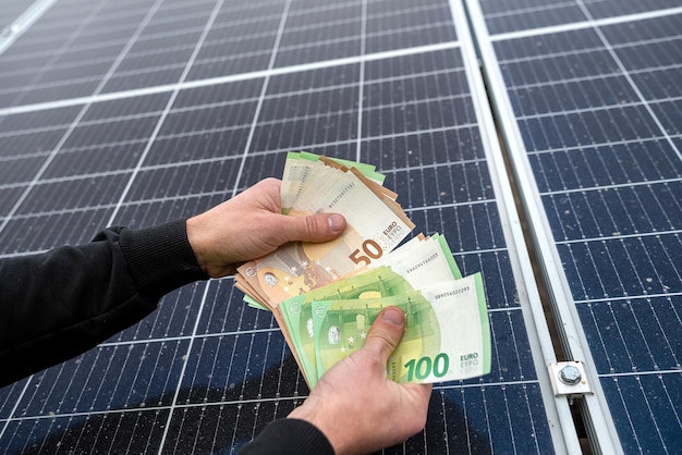 Mooie jonge goed verzorgde handen houden een grote som euro's in de buurt van de geïnstalleerde zonnepanelen. Groene stroom concept