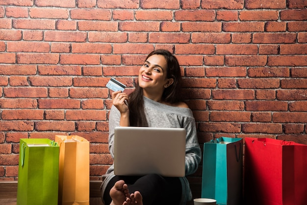 Mooie jonge Indiase vrouw of meisje winkelen met bankpas of creditcard en laptop terwijl ze over een houten vloer tegen een rode bakstenen muur zitten