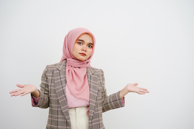 Foto mooie jonge islamitische zakenvrouw met roze hijab, schouders opzuigen, verward kijken, geen idee hebben, staan ondervraagd en onbewust, ik weet niet waarom, handen uitgestrekt, geïsoleerd op een witte achtergrond.