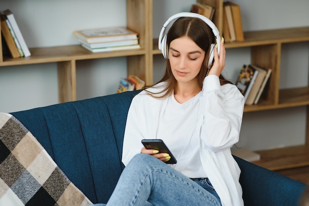 Mooie jonge kaukasische vrouw in vrijetijdskleding die van muziek geniet en glimlacht terwijl ze thuis rust jonge vrouw met koptelefoon die thuis laptop gebruikt