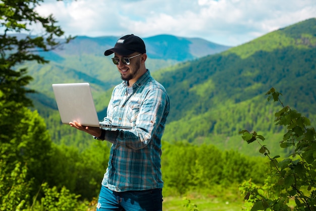 Mooie jonge kerel glimlachend en bezig met laptop in de bergen horizontale foto