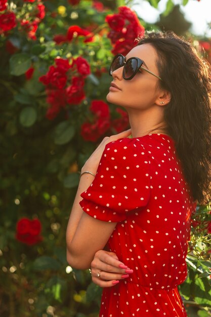 Mooie jonge krullende vrouw met modieuze zonnebril in een rode jurk staat in de buurt van een struik met rode rozen