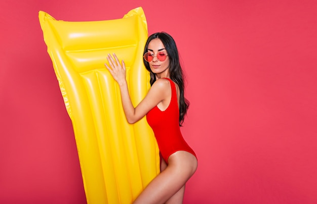 Mooie jonge lachende vrouw gekleed in rode zwembroek en bril poseren met gele opblaasbare matras geïsoleerd over roze achtergrond
