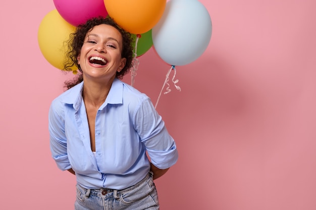 Mooie jonge Latijns-Amerikaanse vrouw met vrolijke en gelukkige emoties lachen camera kijken, ballonnen achter haar rug houden, geïsoleerd op roze gekleurde achtergrond met kopie ruimte voor reclame