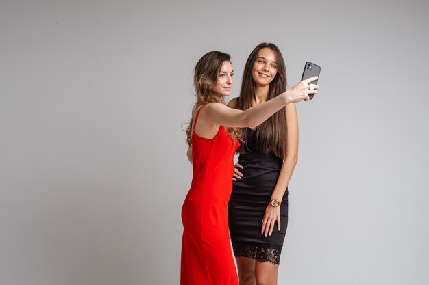 Mooie jonge meisjesbloggers die stijlvolle jurken dragen, maken selfie-fotografie met behulp van de telefoon die zich voordeed op een grijze muur