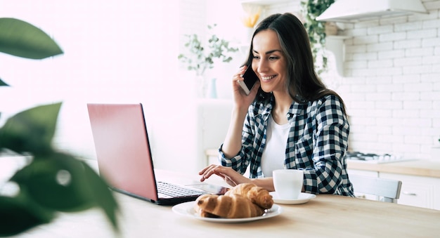 Mooie jonge moderne casual vrouw gebruikt laptop en telefoon op de keuken thuis. Online werken. freelancer