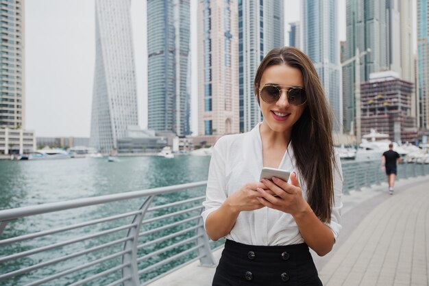 Mooie jonge moderne zakenvrouw permanent in een Dubai Marine en kijken naar haar telefoon.