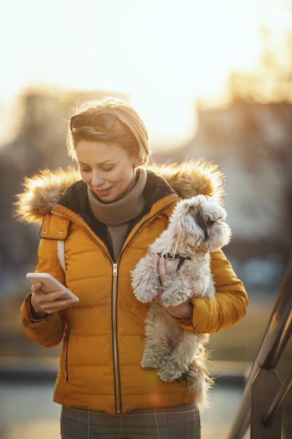 Mooie jonge modevrouw brengt tijd door met haar schattige hondje in de stadsstraat en kijkt naar de smartphone.