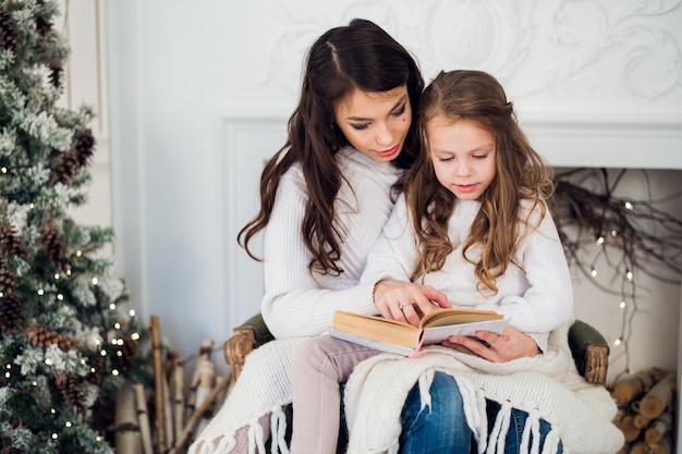 Mooie jonge moeder die een boek leest aan haar schattige dochter in de buurt van boom binnenshuis