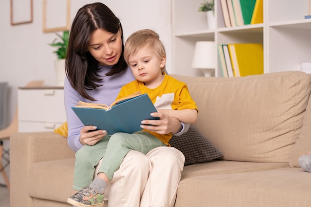 Mooie jonge moeder die haar zoontje met sprookjesboek leest terwijl ze hem op de knieën houdt tijdens thuisrust of zelfisolatie