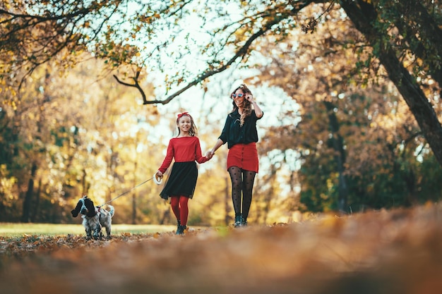 Mooie jonge moeder en haar gelukkige dochter wandelen en hebben plezier in het bos met hun hond bij zonsondergang. Ze houden elkaars hand vast en lachen.