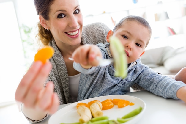 Foto mooie jonge moeder en haar zoon eten thuis fruit.