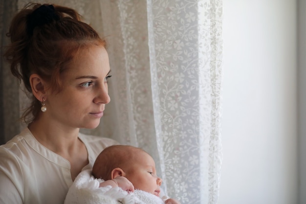 Mooie jonge moeder en pasgeboren baby in haar armen staan bij het raam