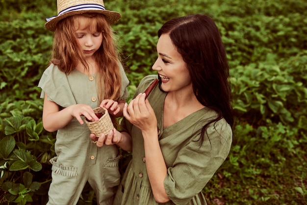 Mooie jonge moeder met haar dochter die pret op een groen gebied met aardbeien heeft