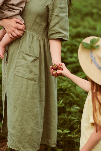 Mooie jonge moeder met haar kinderen in een linnen jurk met een mandje aardbeien verzamelt een nieuwe oogst en heeft plezier met de kinderen